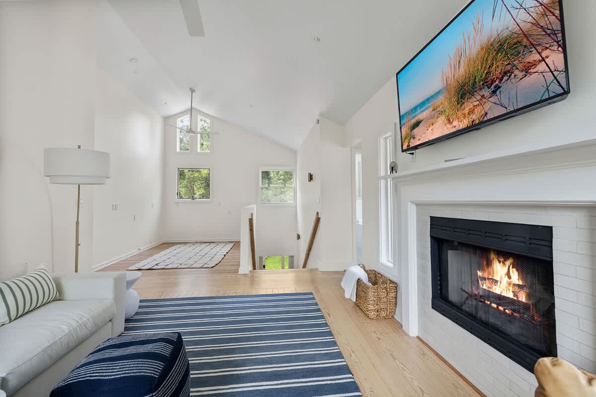Bright living room with light hardwood floors, a white sofa with striped pillows, and a blue and white striped rug. A wall-mounted TV is above a white fireplace with a burning fire, and a floor lamp stands next to the sofa.