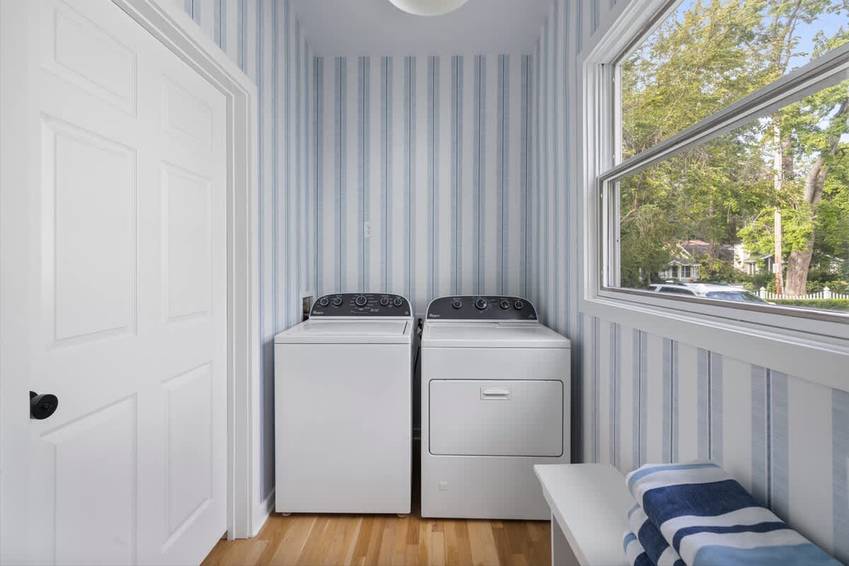 Laundry room with light hardwood floors and blue and white striped wallpaper. A white top-loading washing machine and a front-loading dryer are positioned next to a window, with a small white bench holding folded towels.