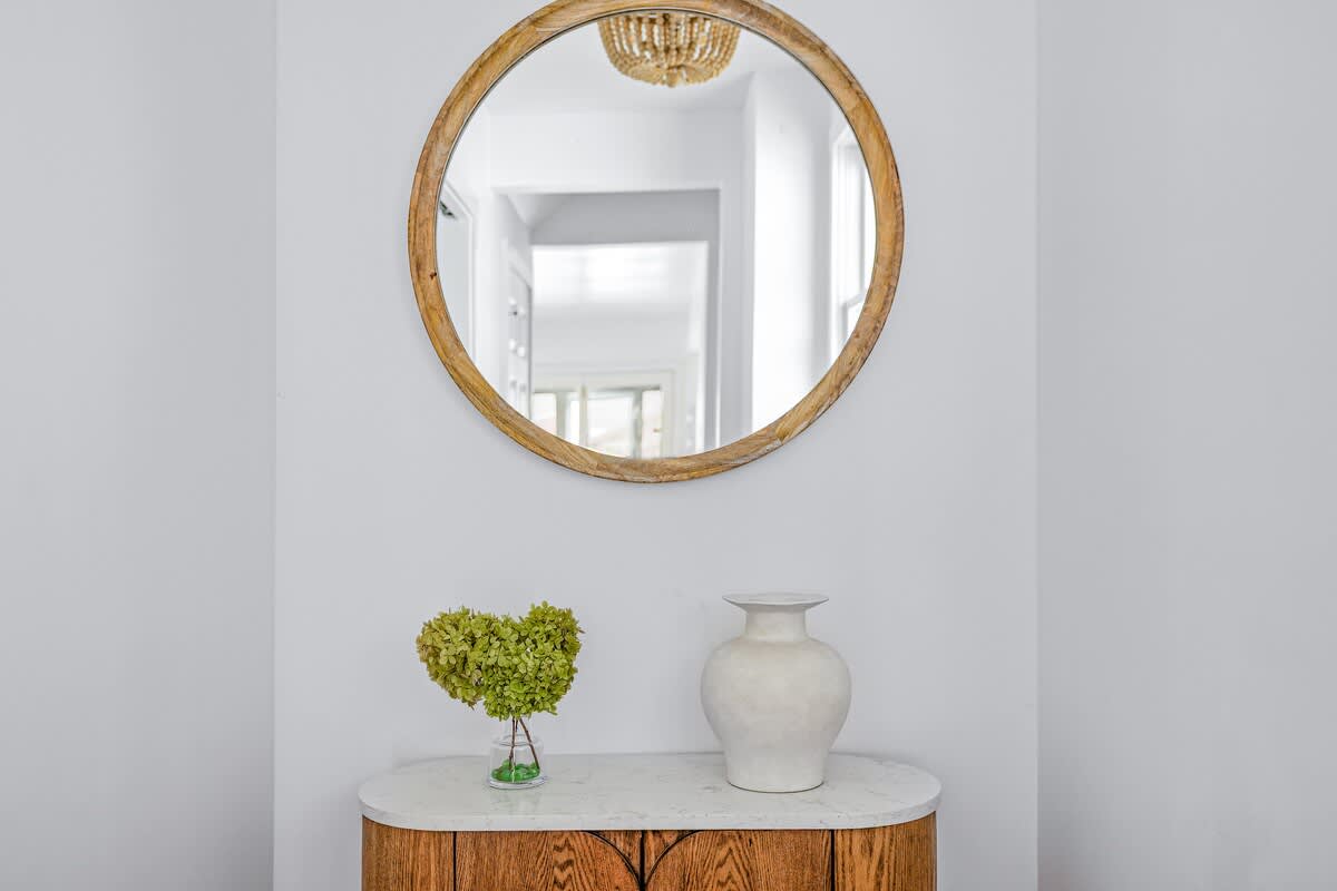 Elegant entryway featuring a large round wooden-framed mirror above a marble-topped console table. A white vase and a heart-shaped plant adorn the table, set against a clean white wall.