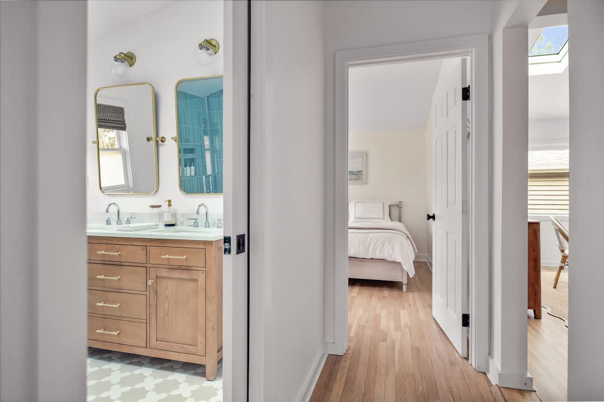 A hallway with light wood flooring leads to a bedroom with a bed visible through an open door, while a glimpse into a bathroom reveals a double vanity with gold mirrors and light blue tiled shower.