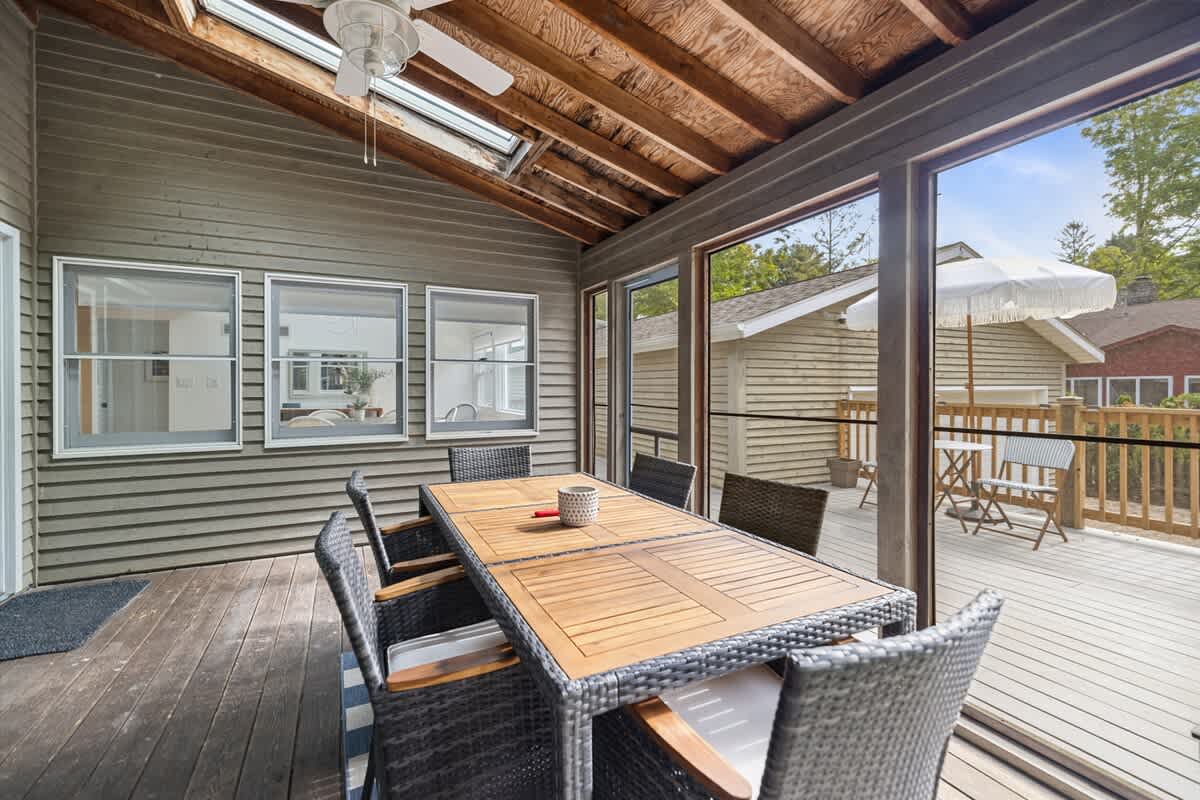 Covered outdoor dining area with a long wooden table and dark wicker chairs, set on a wooden deck. Large windows and a skylight provide natural light, with views of the surrounding property and a ceiling fan overhead.