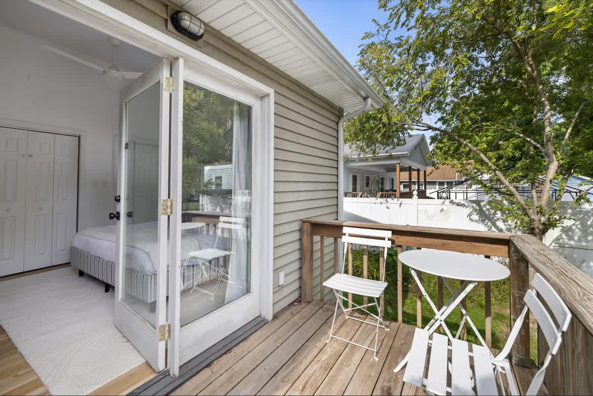 A cozy wooden balcony extends from a bedroom through glass French doors, furnished with a white metal bistro set, offering views of neighboring houses and lush trees.