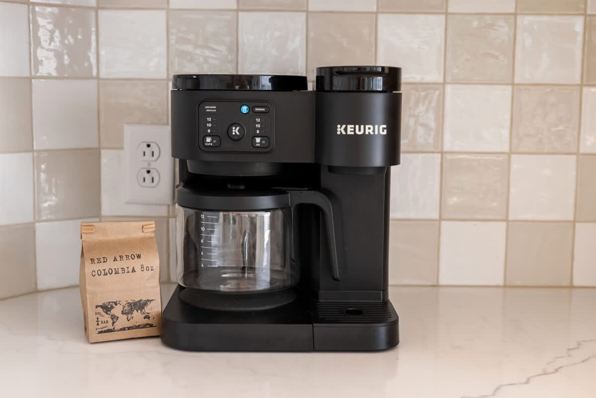 A black Keurig coffee maker sits on a white countertop against a white checkered tile backsplash, next to a brown bag of "Red Arrow Colombia" coffee beans. A white electrical outlet is visible on the wall.