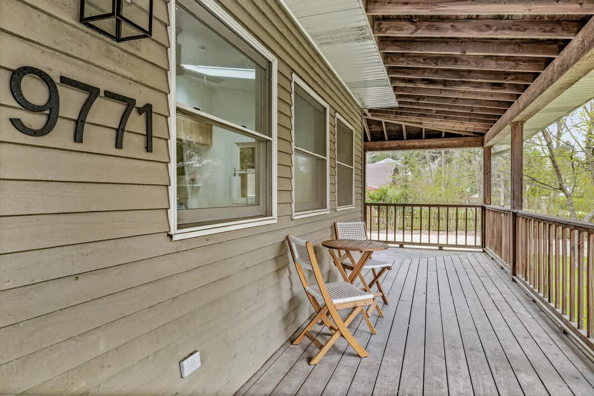 A long, covered wooden porch with a railing and two wooden chairs and a small table, featuring large black house numbers "9771" on the light brown siding of the house.
