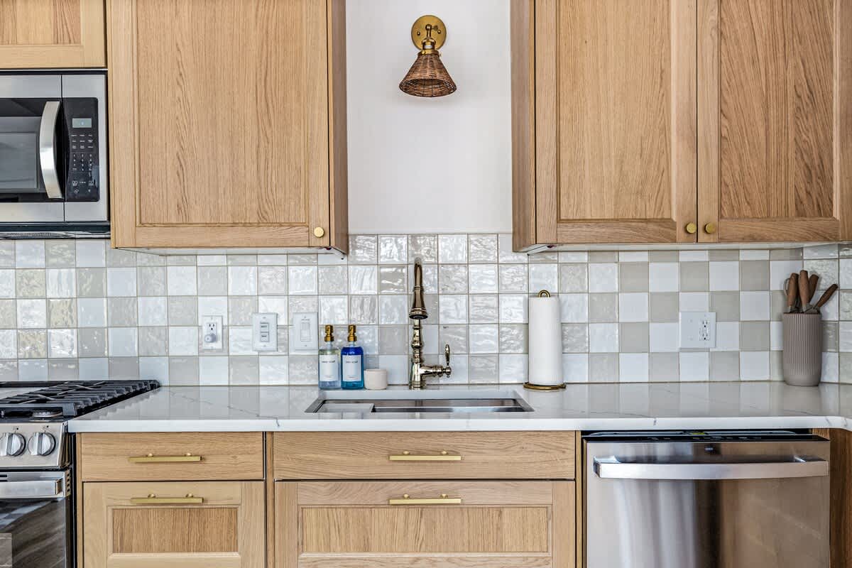 Close-up of a modern kitchen counter with a stainless steel sink, brass faucet, and white checkered tile backsplash. Light wood cabinets and drawers with brass pulls are visible, along with a microwave and a woven wall sconce.