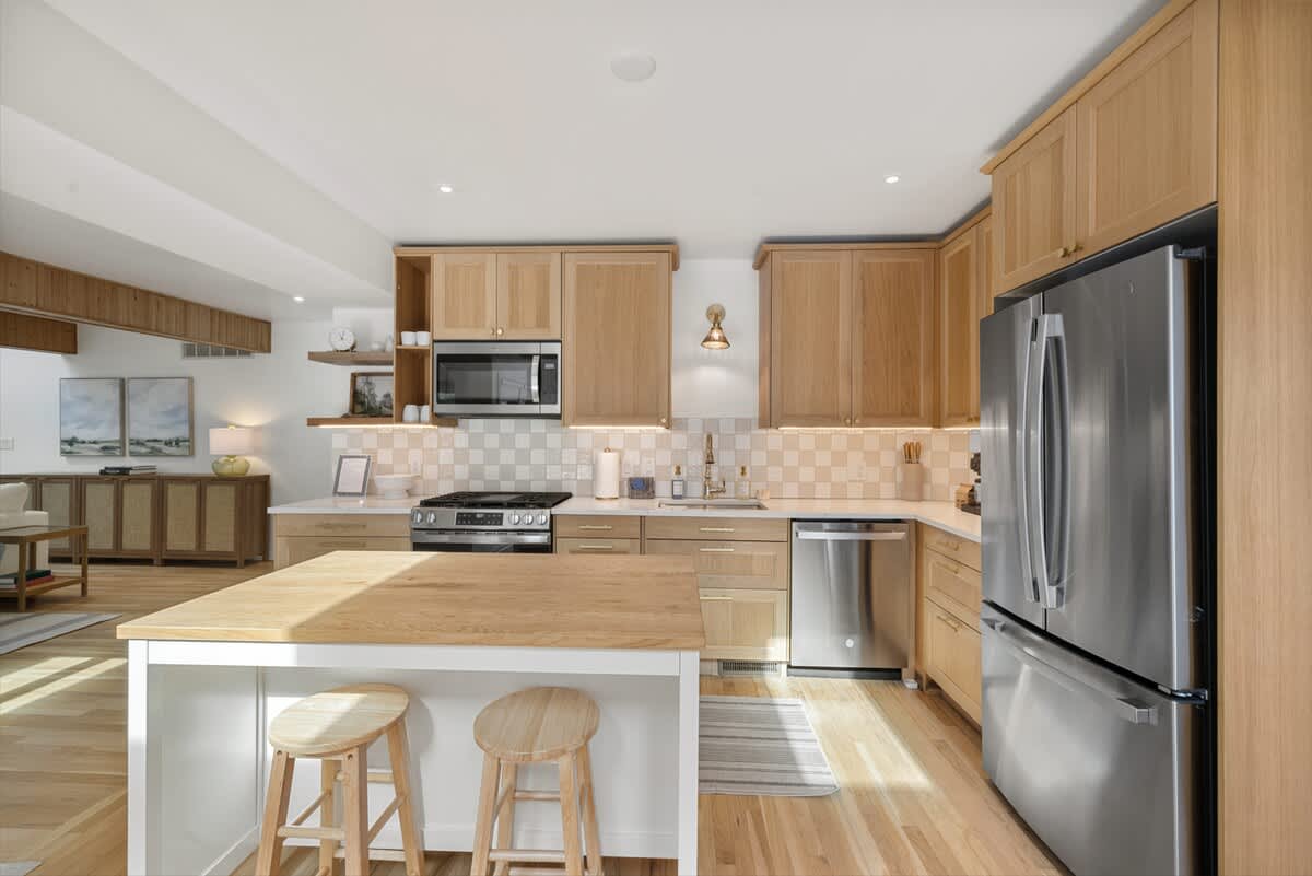 Bright, open-concept kitchen with light wood cabinetry, stainless steel appliances, a white checkered tile backsplash, and a large wooden island with two stools. Hardwood floors reflect natural light from overhead recessed lighting.