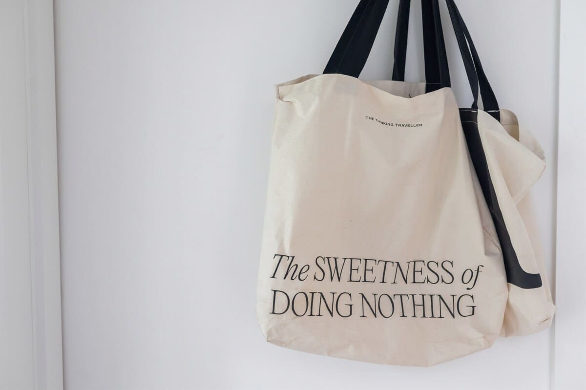 Close-up shot of a cream-colored canvas tote bag with black handles, hanging against a white wall. The bag features the text "The Sweetness of DOING NOTHING" in black lettering.
