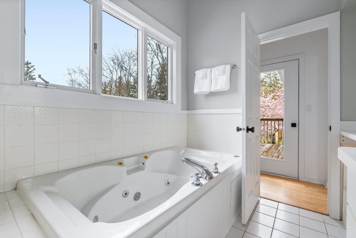 Bathroom featuring a large white jetted bathtub with chrome fixtures, surrounded by white subway tile wainscoting. White square floor tiles and a large window offering a view of trees complete the space.