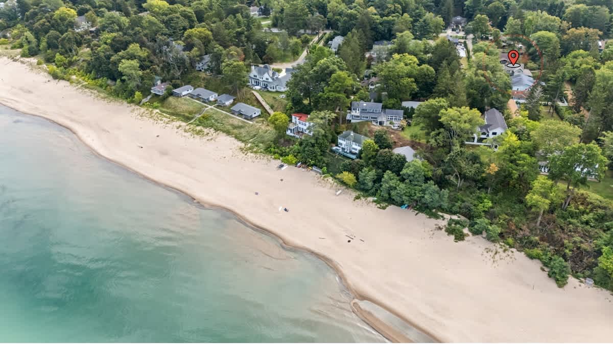 Aerial view showcasing a sandy beach coastline next to a turquoise body of water, with several houses nestled among dense green trees. A red pin highlights the property's location near the beach.