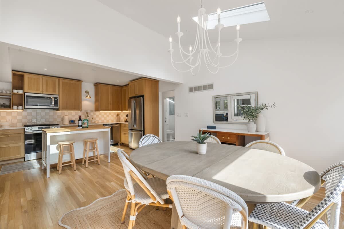 Wide view of the open-concept dining and kitchen area, showcasing a light wood oval dining table with wicker chairs, a white chandelier, and a modern kitchen with light wood cabinetry and stainless steel appliances. Natural light streams in from large windows and a skylight.
