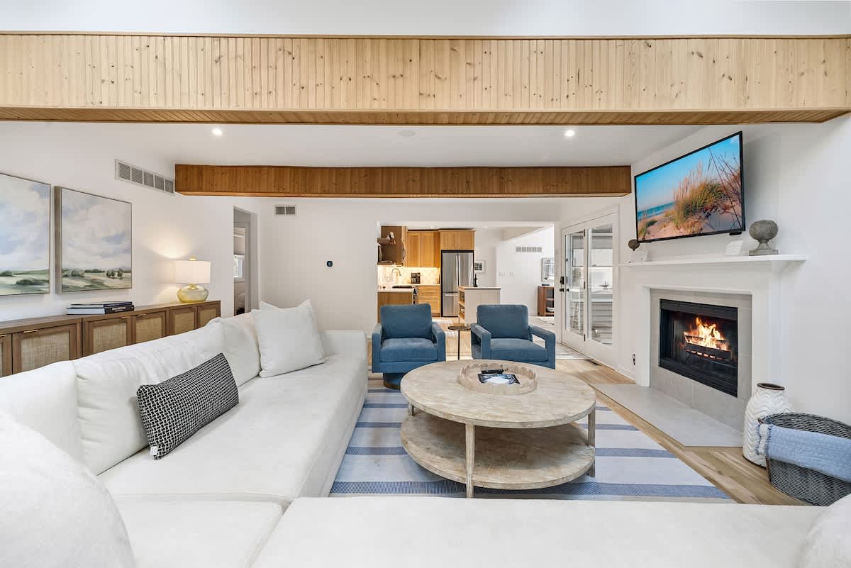 Wide view of the living room, showing a large white sectional sofa, a round wooden coffee table, and a striped rug. The fireplace with a TV above it is visible on the right, and the kitchen can be seen in the background.