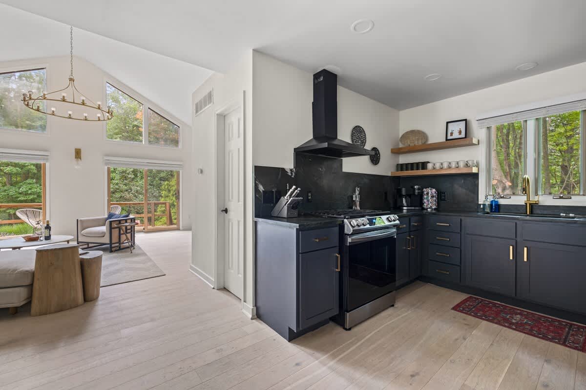 An open-concept living space features a modern kitchen with dark cabinetry, black countertops, and a black range hood, flowing into a bright living area with vaulted ceilings, large windows, and light wood floors.