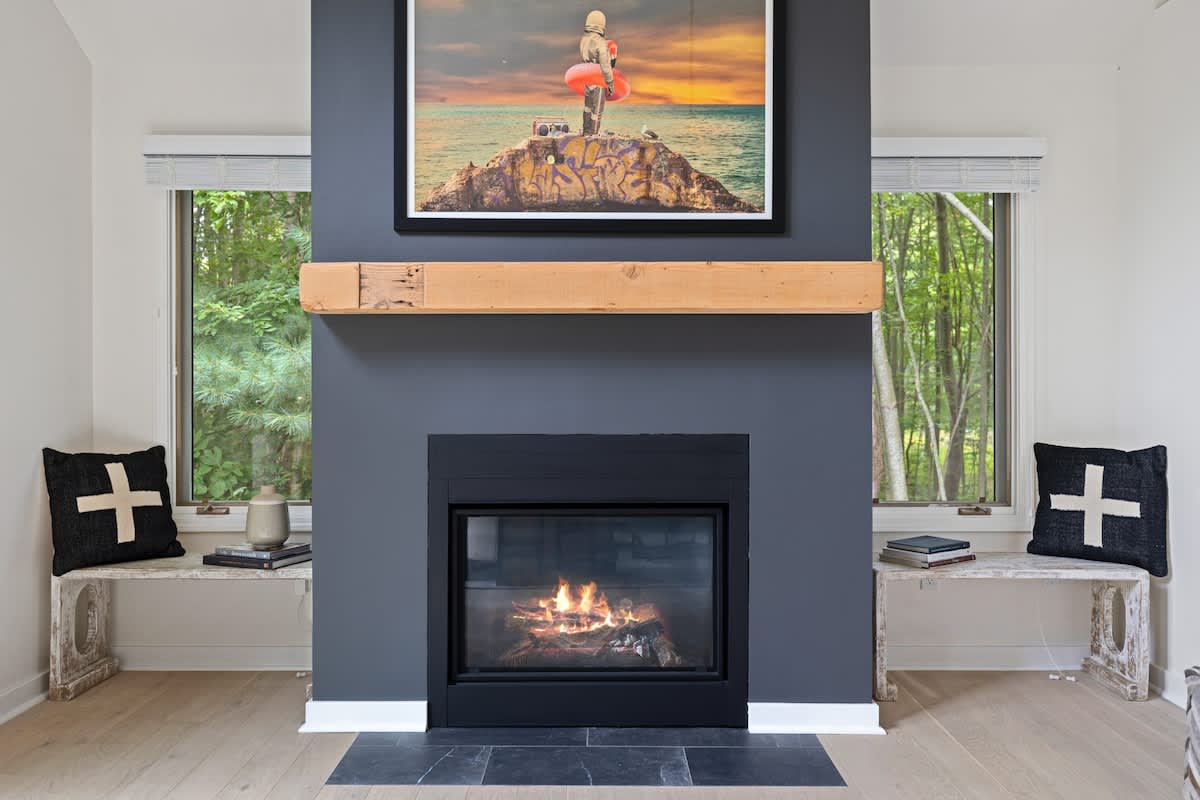 A modern fireplace with a black insert and dark gray surround is topped by a rustic wooden mantel and contemporary artwork, flanked by two windows and white benches with decorative pillows.