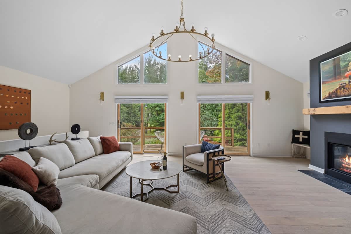 A spacious living room with a vaulted ceiling and large windows, including triangular transom windows, offering views of lush greenery, featuring a light sectional sofa, a round coffee table, and a dark gray fireplace with a wooden mantel.