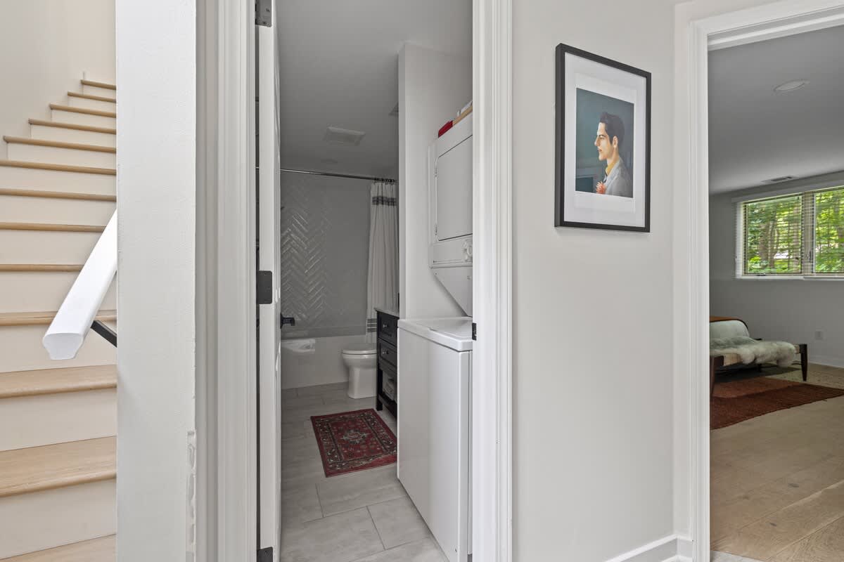 A narrow hallway leads to a bathroom with a white tiled shower/tub combo, and a stacked white washer and dryer unit is visible on the right, next to a framed portrait.