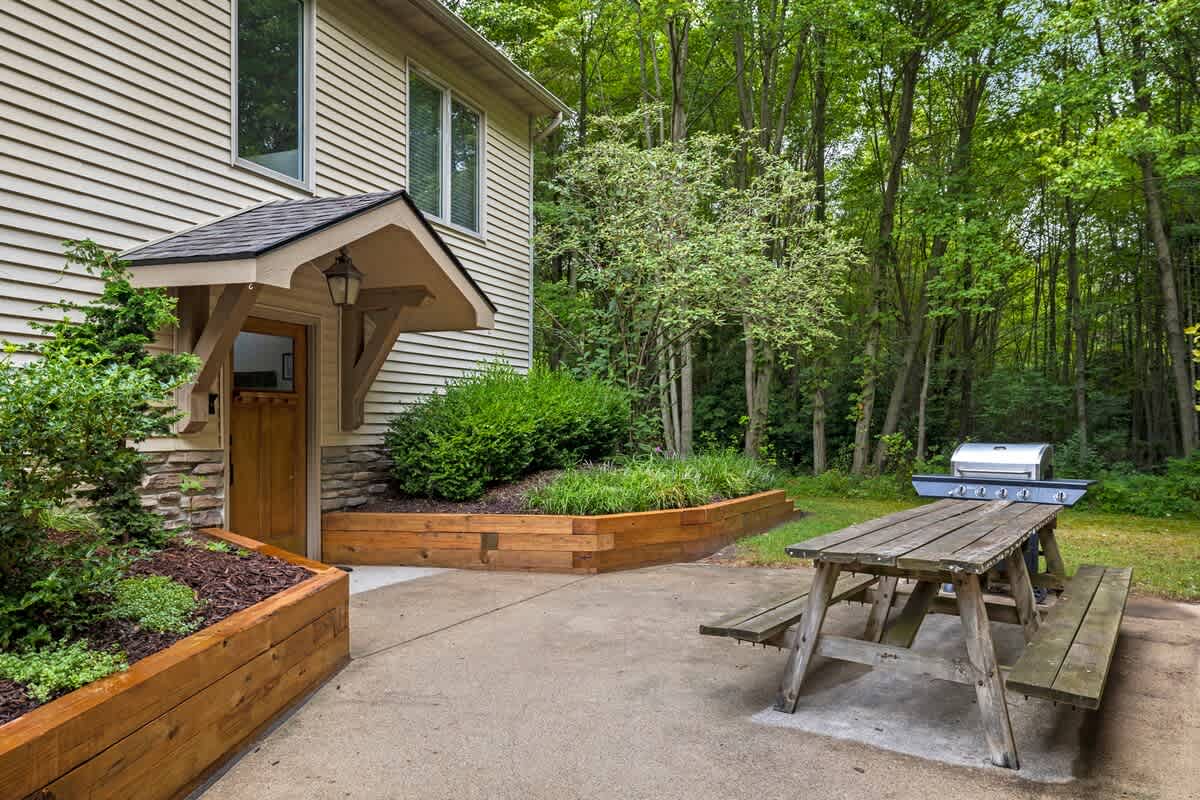 The side entrance of a light-sided house with a covered entryway, surrounded by landscaped garden beds, and a concrete patio with a rustic wooden picnic table and a BBQ grill.