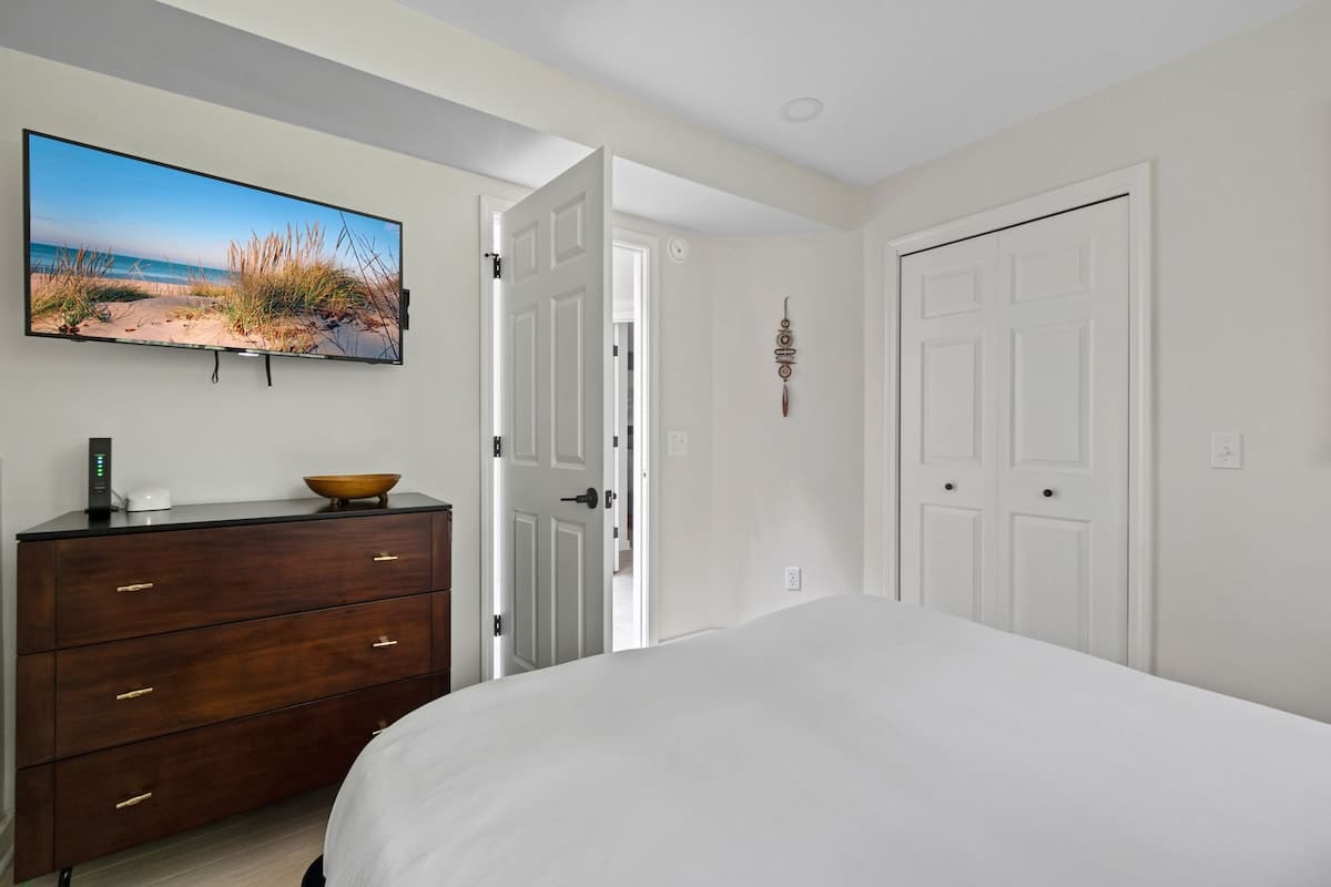 A bright and modern bedroom featuring a large wall-mounted TV above a dark wooden dresser with gold handles, a white bed with crisp white bedding, and light hardwood floors.