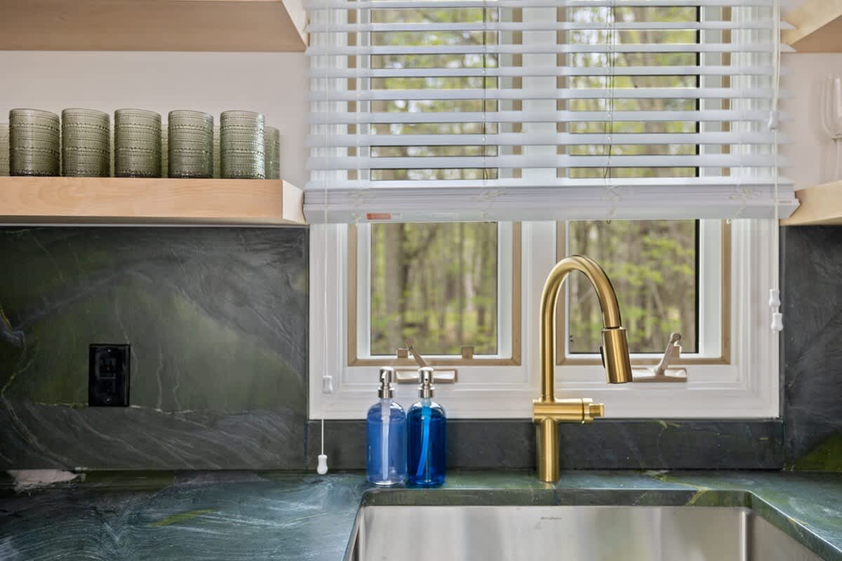 A close-up of the kitchen sink area shows a gold gooseneck faucet, a stainless steel sink, and blue soap dispensers against a dark green marble-patterned backsplash and countertop, with a window overlooking trees.
