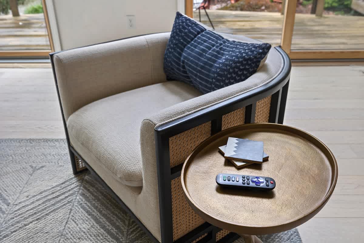 A detailed shot of a stylish armchair with a cane back and black frame, upholstered in a light fabric, with a blue patterned throw pillow, next to a small round brass side table holding a remote control and coasters.
