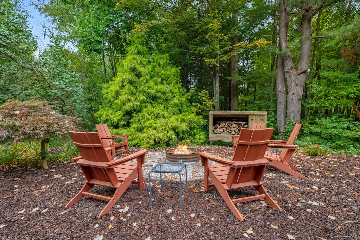 A cozy outdoor fire pit area features a round metal fire pit surrounded by four rustic wooden Adirondack chairs, set on a bed of wood chips. A wooden storage unit filled with firewood and dense green foliage are in the background.