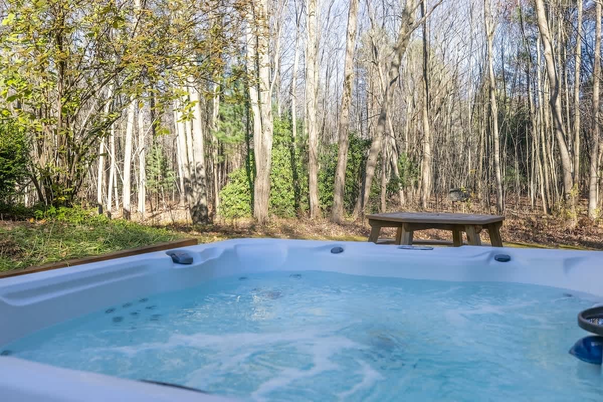 A white hot tub with bubbling water is set in a serene wooded environment, surrounded by tall, bare trees and some evergreens, with a small wooden bench visible in the background.