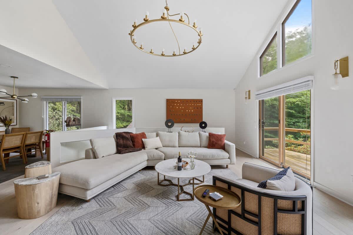 A bright living room with a large light-colored sectional sofa, a round marble coffee table, and a cane-backed armchair, all on a geometric rug, with large windows and a sliding glass door leading to an outdoor deck.