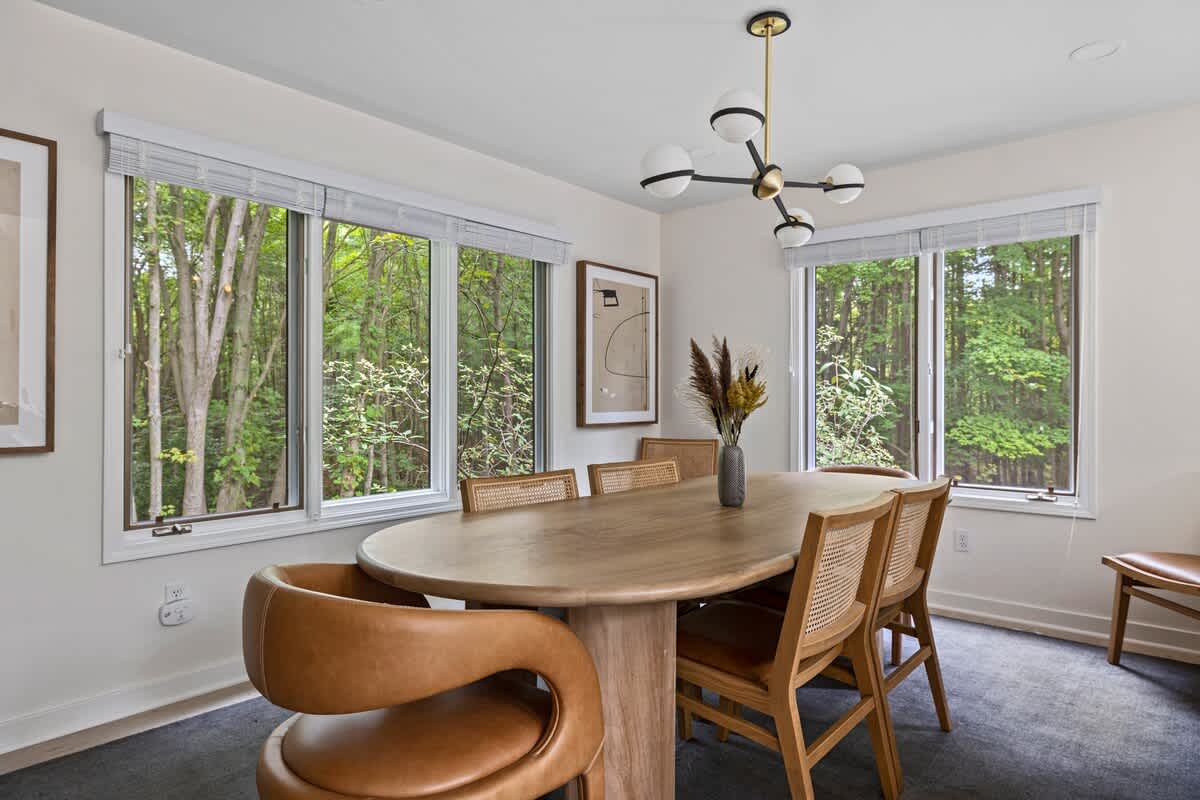 A close-up of the dining area featuring an oval wooden table with a centerpiece vase, surrounded by cane-backed and curved leather dining chairs, with multiple windows providing views of dense green trees.