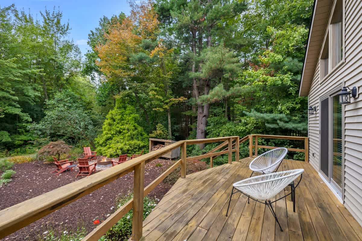 A spacious wooden deck with two modern white chairs, overlooking a fire pit area furnished with Adirondack chairs, all surrounded by lush green trees and natural landscaping.