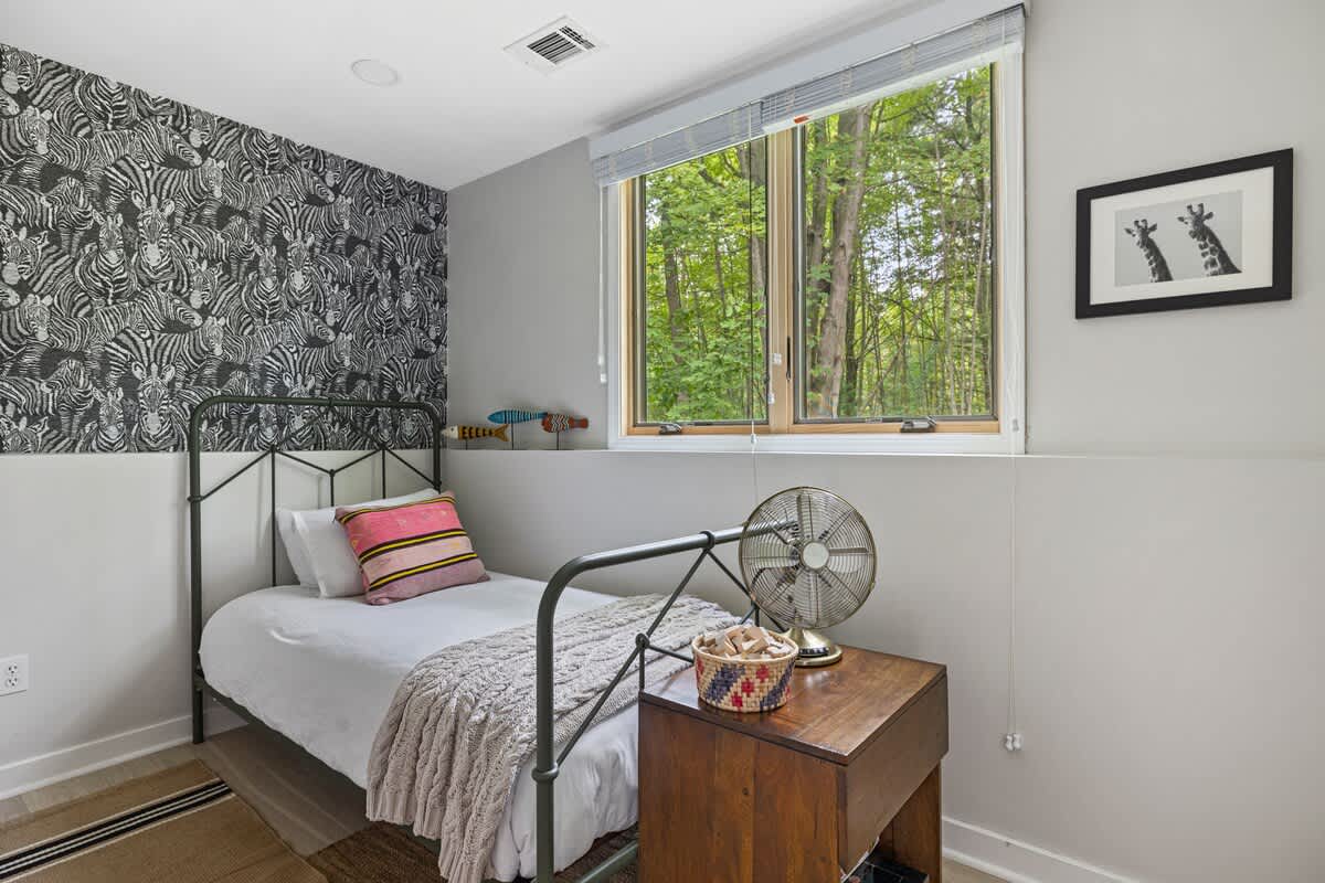 A single metal-frame bed with a colorful pillow and knitted throw, positioned next to a wooden nightstand with a vintage fan, against a gray wall with a zebra print accent.