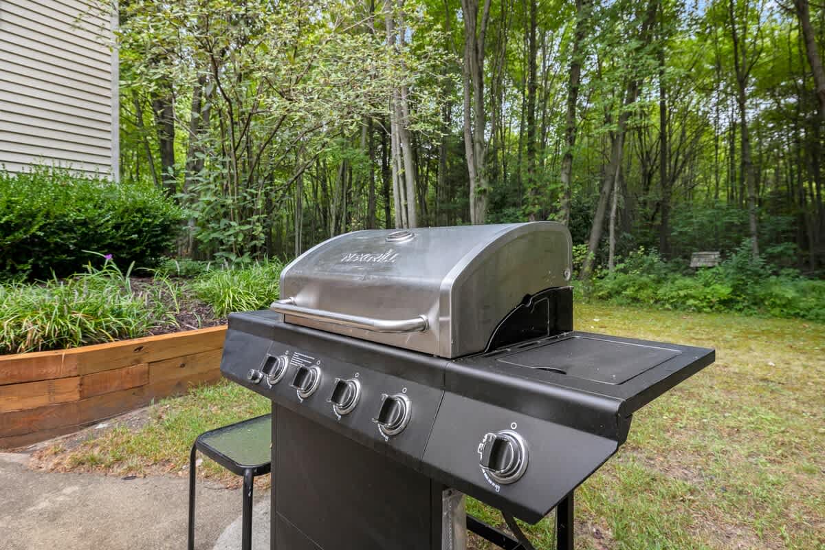 A large stainless steel and black gas BBQ grill stands on a paved area next to a raised wooden garden bed, with a vibrant green lawn and a dense forest of trees in the background under a bright sky.