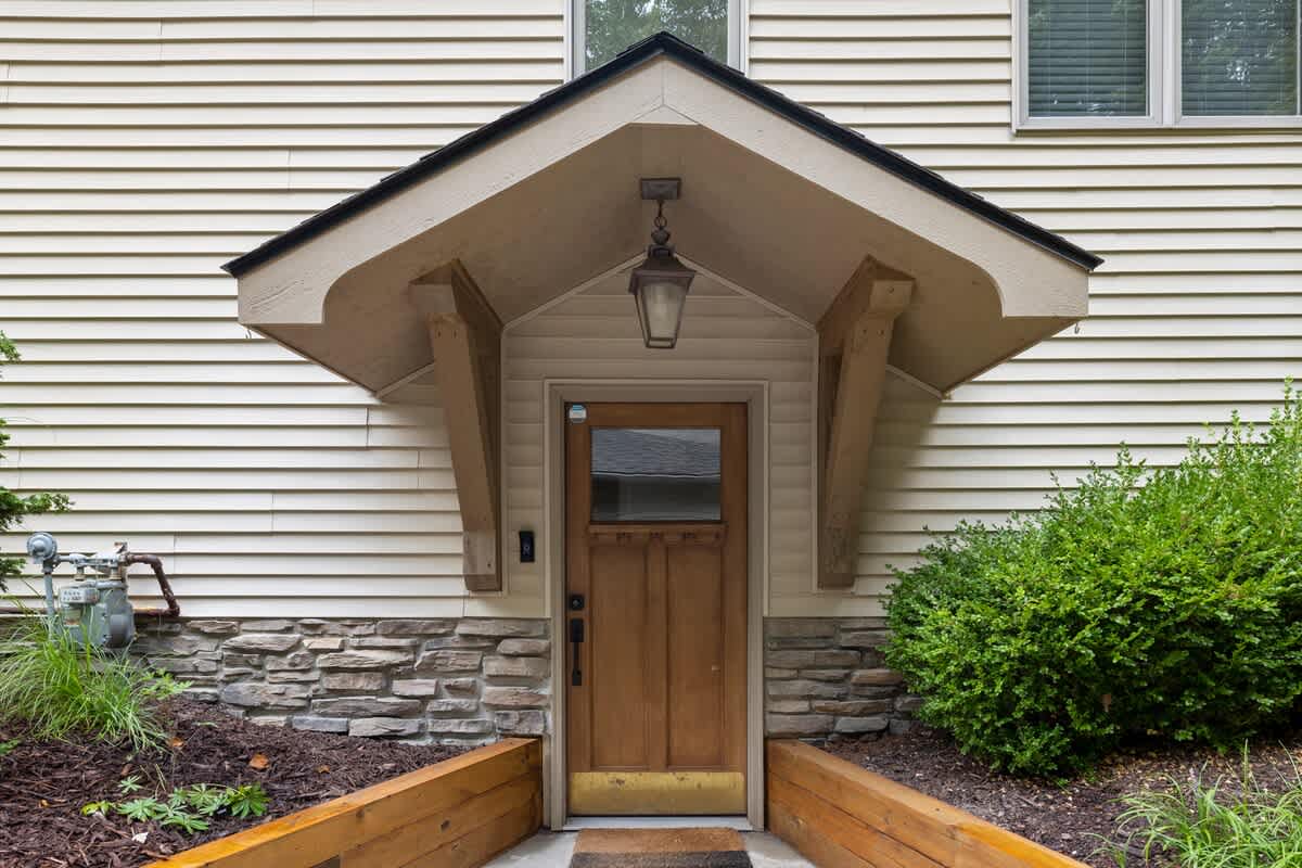A close-up of the house's front entrance features a wooden door with a small window, sheltered by a covered porch with decorative brackets and an overhead lantern, set against light siding and a stone foundation.