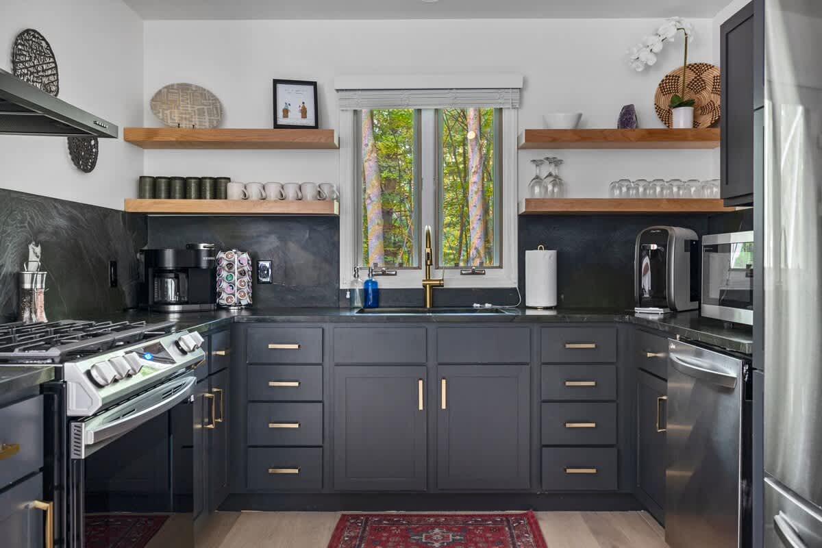 A modern kitchen with dark gray base cabinets and black countertops, featuring a gas range, a gold-toned faucet over a sink, open wooden shelving, and a window looking out to green trees.