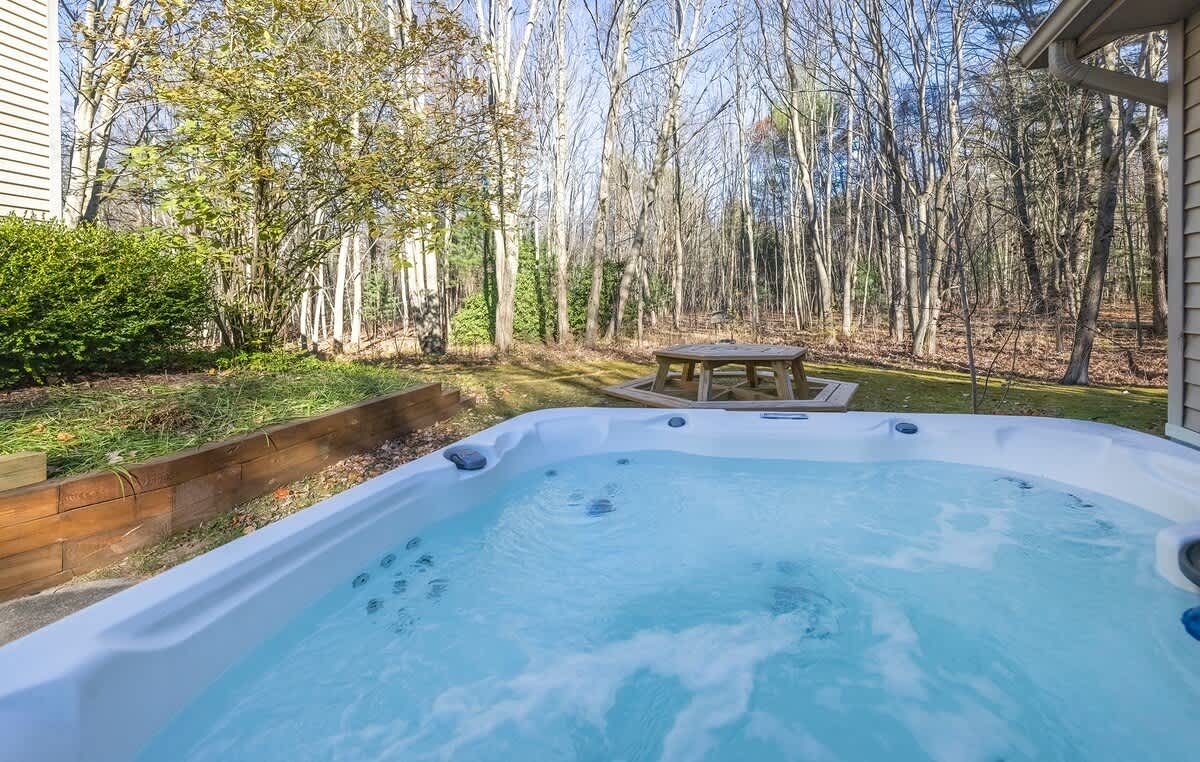 A large white hot tub with bubbling water is situated on a concrete patio, overlooking a lush green lawn and a dense forest of tall, bare trees. A wooden picnic table is visible further back in the yard.