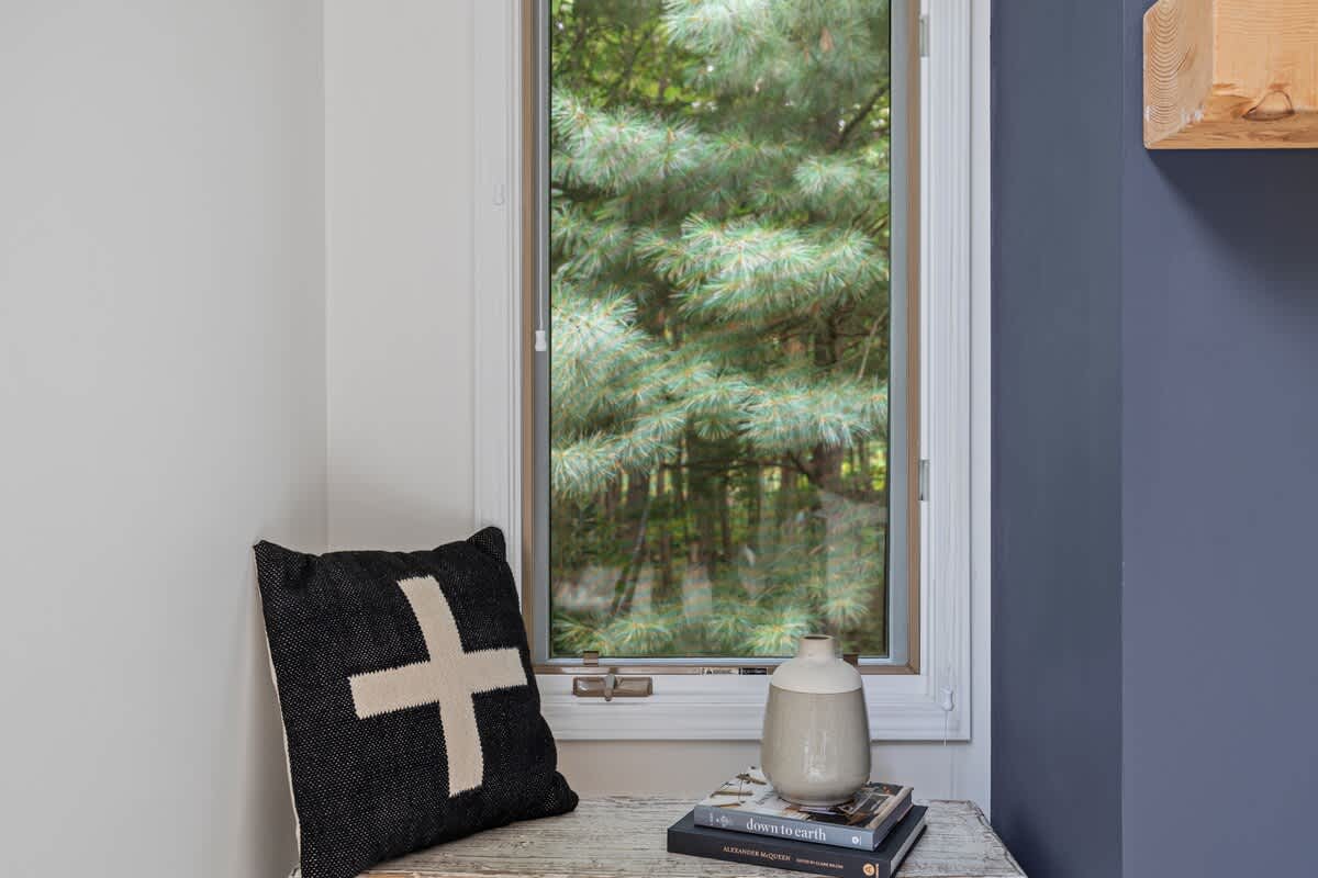 A close-up of a cozy corner next to a window, featuring a black and white patterned throw pillow on a rustic wooden bench, with a ceramic vase and books, offering a glimpse of green trees outside.