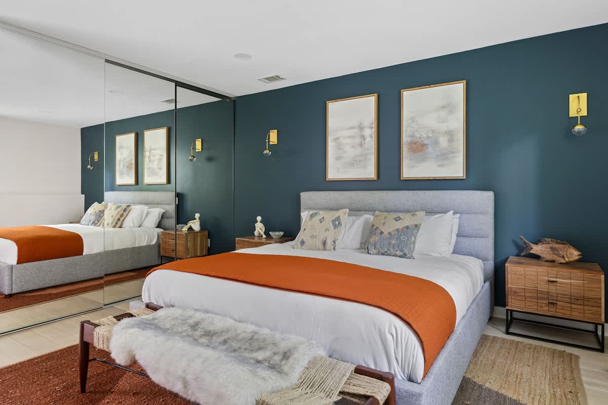 This view of the master bedroom highlights a large mirrored closet reflecting the king-sized bed with its grey frame, orange throw, and dark teal accent wall, creating a sense of expanded space.