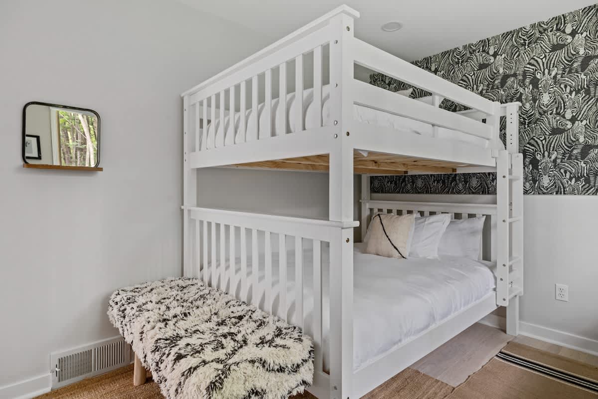 A children's bedroom featuring a sturdy white wooden bunk bed with white bedding, a striking black and white zebra print accent wall, and a textured bench with a shaggy throw.