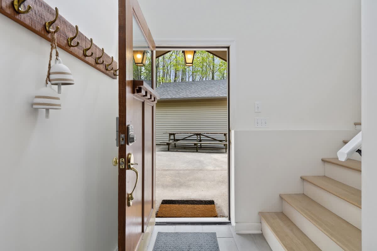 The interior entryway shows a wooden front door slightly ajar, revealing an outdoor concrete area and a picnic table, with a wooden coat rack on the wall and light-colored stairs leading up.