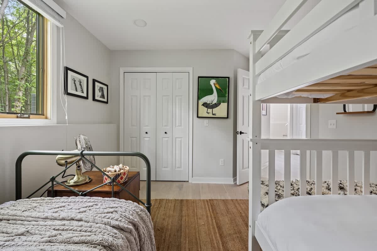 A bright bedroom with a white wooden bunk bed and a single metal-frame bed, featuring white bi-fold closet doors and a window offering views of lush green trees.