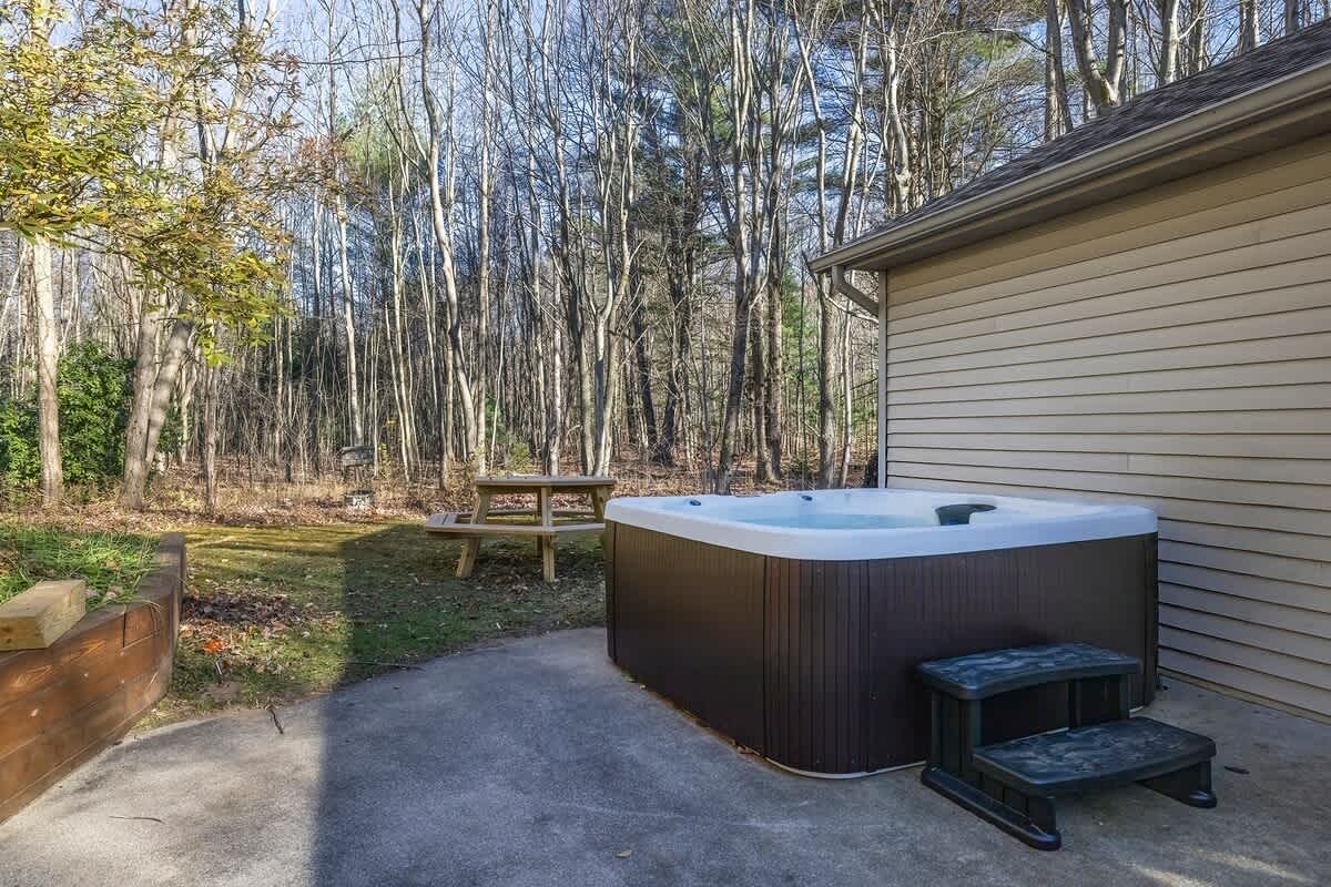 A modern hot tub with steps situated on a concrete patio next to the house, surrounded by a grassy yard and a dense forest of bare trees in the background.
