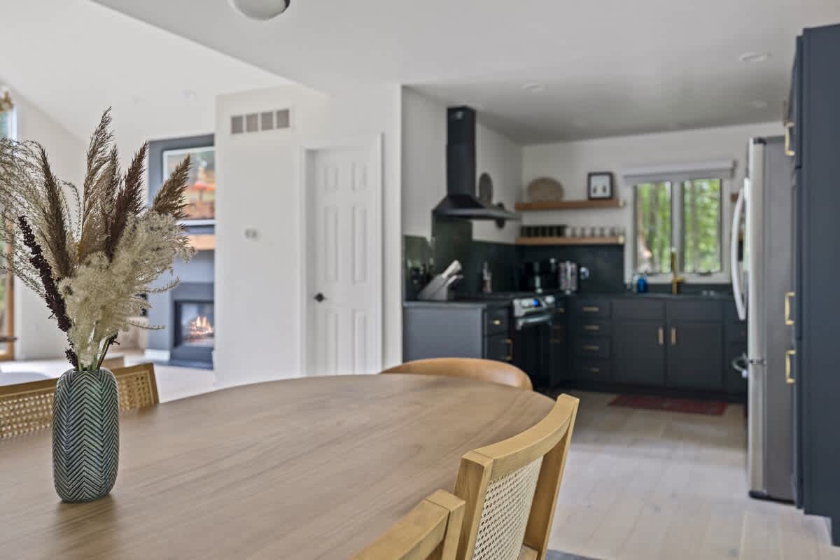 A light wood dining table with cane-back chairs is in the foreground, looking towards an open-concept kitchen with dark cabinets and a living area with a fireplace visible in the distance.
