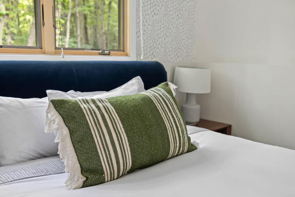 A close-up of the bed in the second bedroom shows a navy upholstered headboard, crisp white bedding, and a decorative green and white striped pillow with fringe, against a patterned accent wall.
