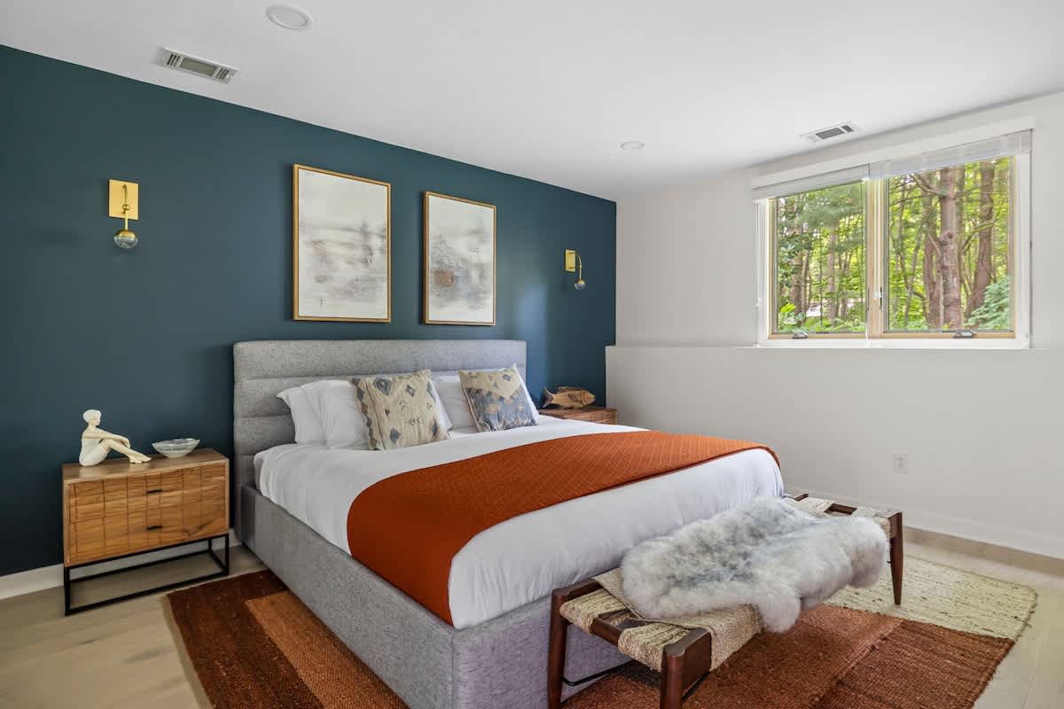 A spacious bedroom features a king-sized bed with a grey upholstered frame, an orange throw, and a dark teal accent wall, flanked by wooden nightstands and gold wall sconces, with a large window offering a view of trees.