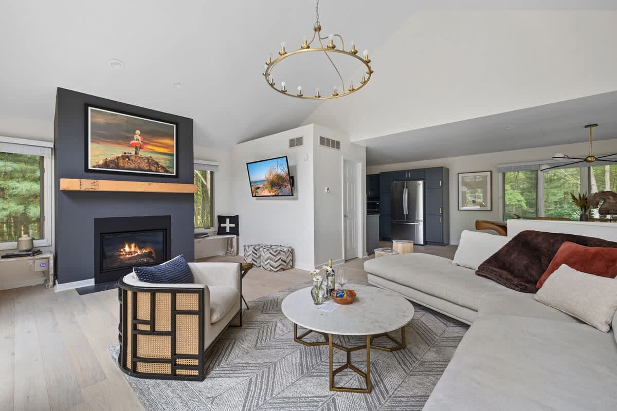 A modern living room with a vaulted ceiling, light wood floors, a large sectional sofa, a round marble coffee table, and a dark gray fireplace with a wooden mantel and a large framed artwork above it.