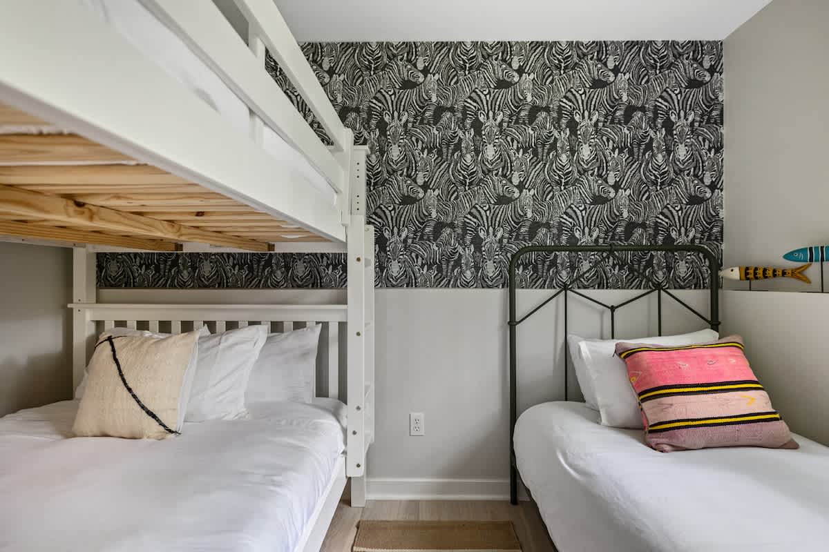 A cozy bedroom featuring a white wooden bunk bed and a single metal-frame bed, both with white bedding, set against a distinctive black and white zebra print accent wall.