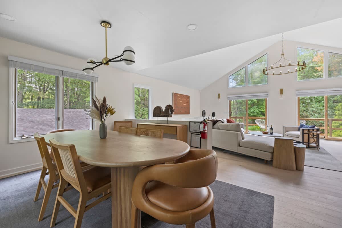 An open-concept view showing the dining area with a large oval wooden table and a mix of cane-backed and leather chairs, adjacent to the living room, with large windows overlooking green trees.
