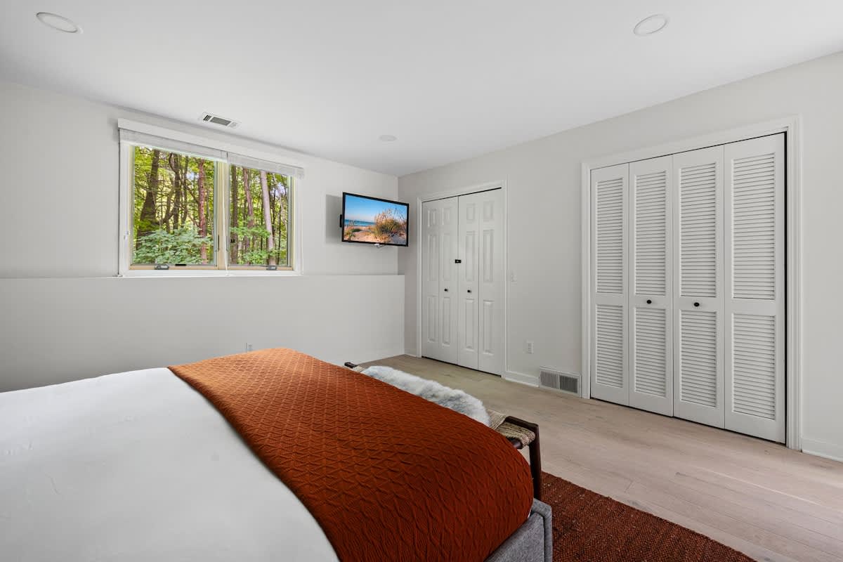 The bedroom features a wall-mounted TV opposite the bed, with two white bifold closet doors and a window providing natural light and a view of the surrounding trees.