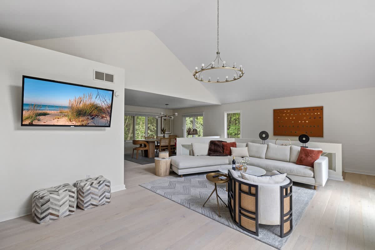 A wide view of the living room featuring a light-colored sectional sofa, a round coffee table, and a cane-backed armchair on a patterned rug, with a wall-mounted TV visible on a white wall and a dining area in the background.