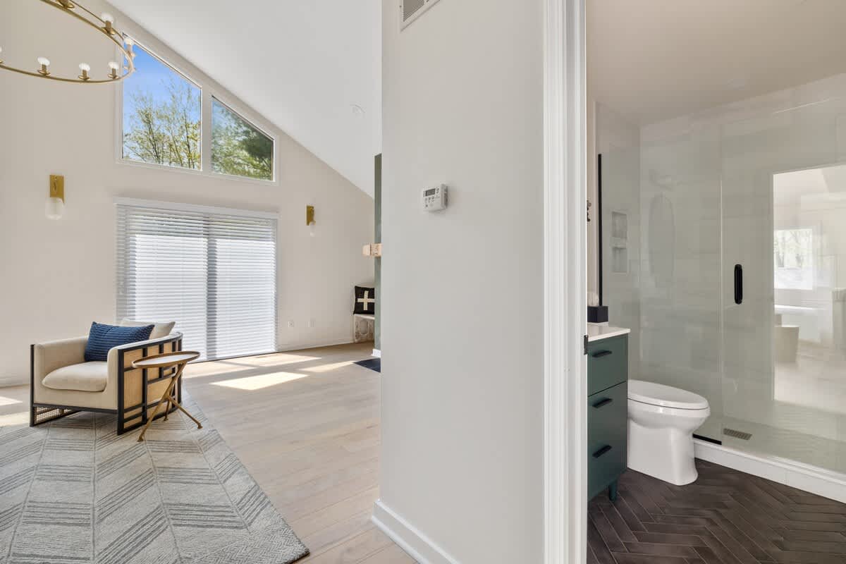 A transitional view from a hallway with light hardwood floors, showcasing a spacious living area with large windows and a modern armchair, and a glimpse into a bathroom with a dark green vanity.