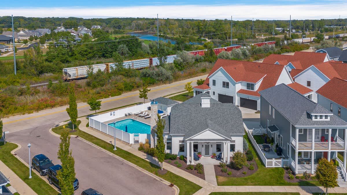 An aerial view of the residential neighborhood, showing several houses with varying roof colors, a community pool area, and a train track running along the edge of the development.