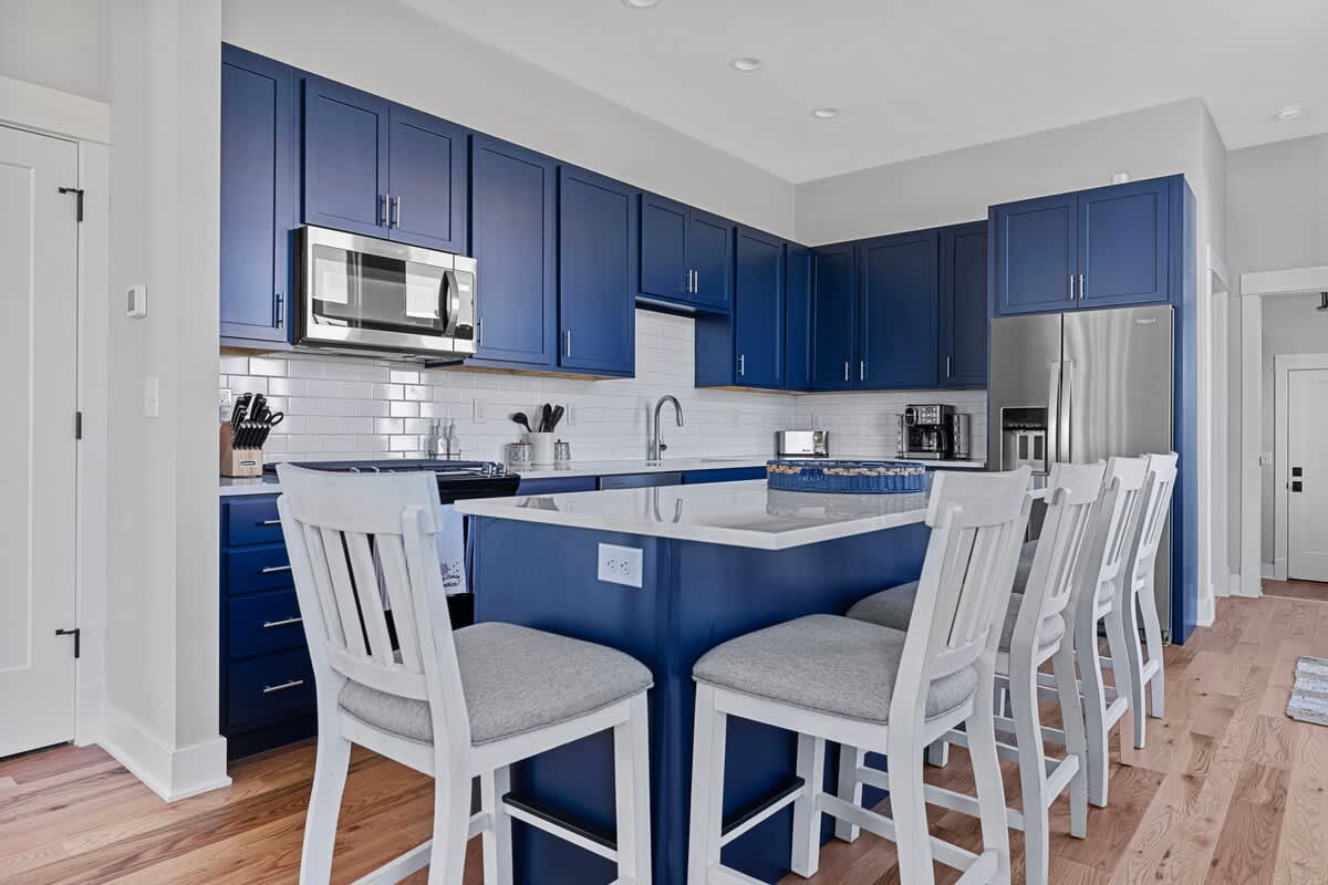 Open-concept kitchen with dark blue shaker cabinets, white countertops, and a large island with seating for four on white bar stools, connecting seamlessly to the living area.
